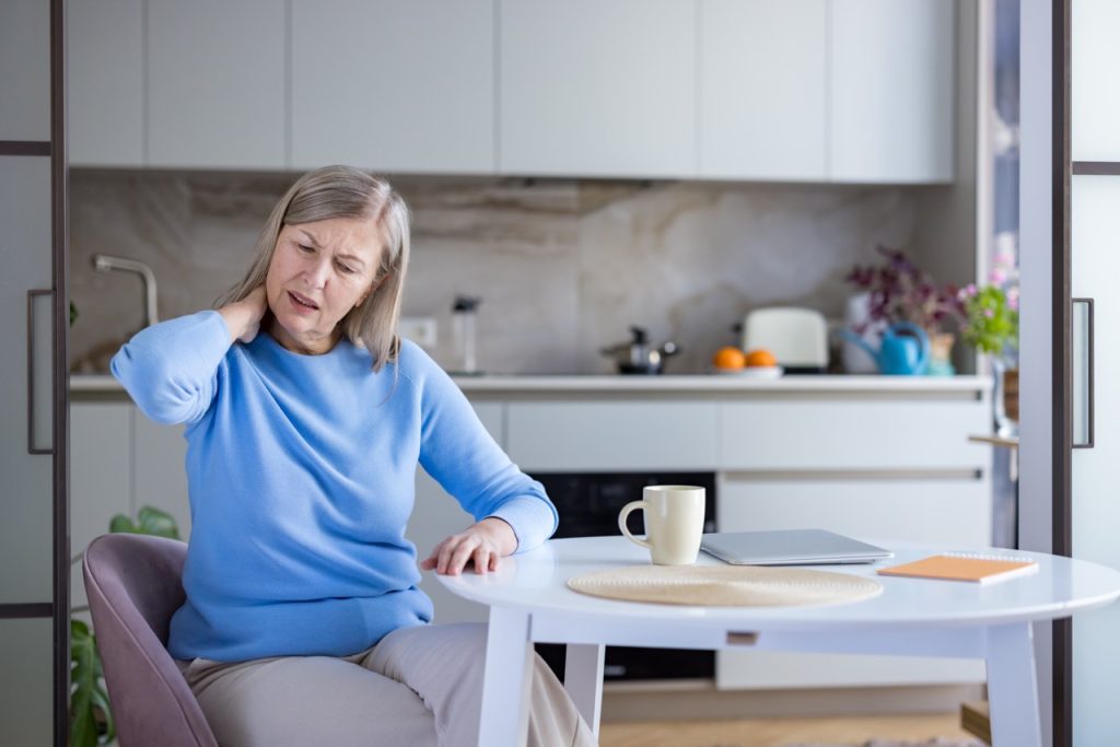 Senior woman experiencing chronic neck pain and stiffness, touching her hurting neck and shoulder while sitting at a kitchen table, symbolizing health issues and physical discomfort in old age