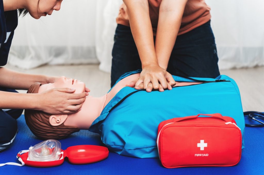 person performing cpr on cpr training dummy in a blue shirt with red first aid kit nearby