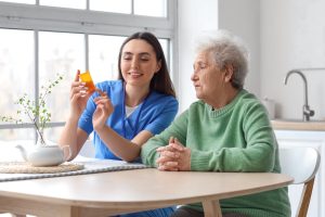 Young TMA caregiver helping senior woman take medication in kitchen