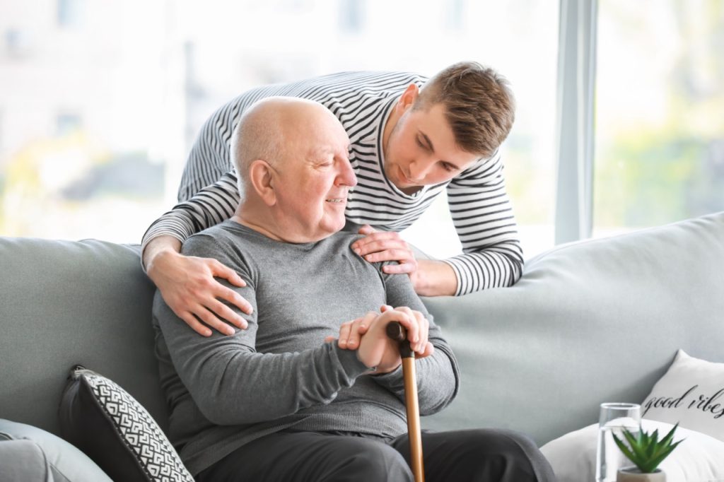 A young man caregiver with a senior man holding a cane and sitting on a couch in a bright room