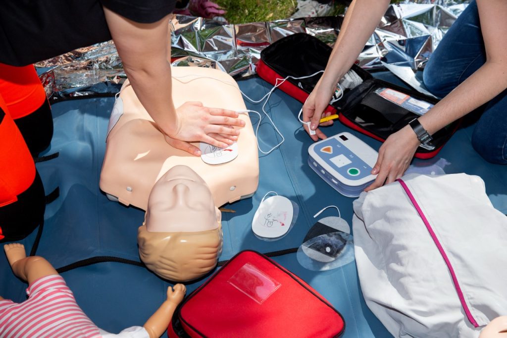 An overhead view of a CPR training session. One person demonstrates chest compressions on an adult mannequin equipped with AED pads, while another person operates a training AED device nearby.