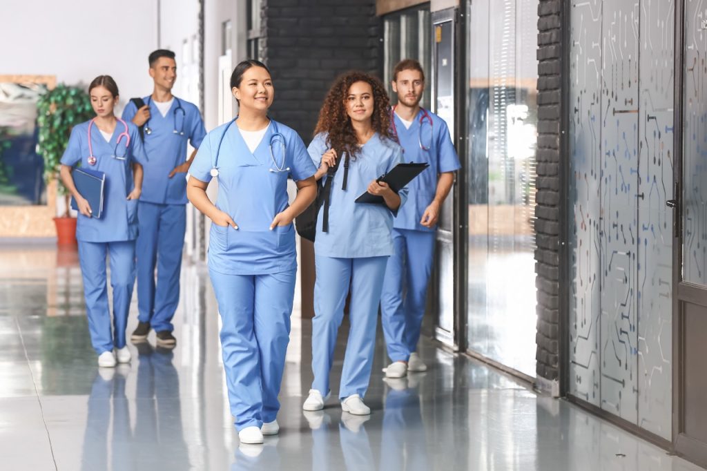 A group of smiling nursing students in blue scrubs walking through a campus hallway, illustrating the transition from a CNA into a nursing school program.